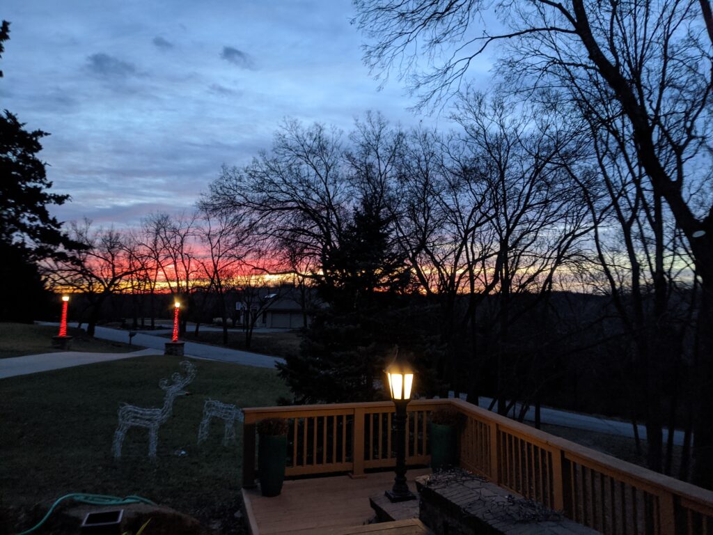 Wooden deck with lamppost at dusk, colorful sunset, bare trees, decorative lit reindeer, red-lit columns.