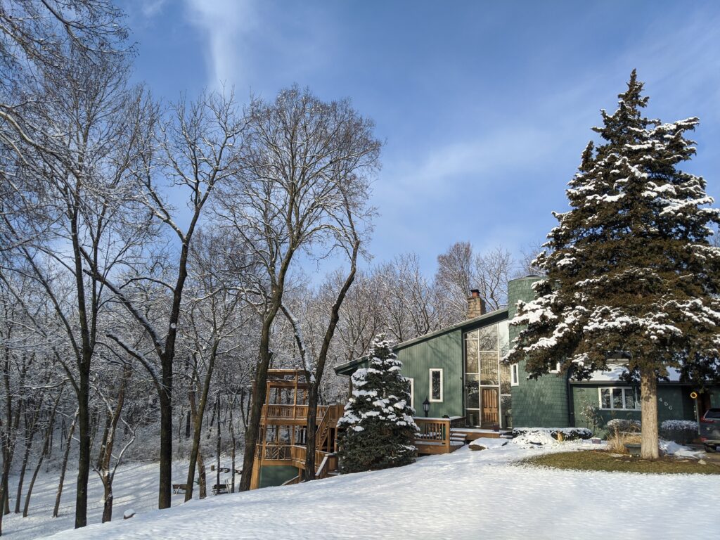 Modern green house with large windows and a deck, surrounded by snowy trees and a clear blue sky.