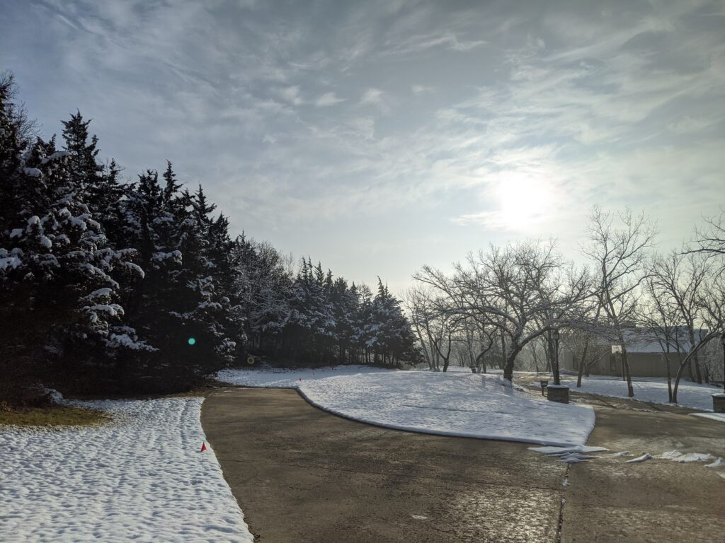 Pathway through snow-covered landscape with evergreen trees on left, bare trees, and building on right.