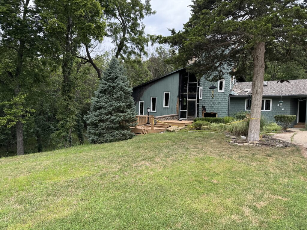 Damaged wooden house with caution tape, well-maintained lawn, mature trees, and various shrubs.