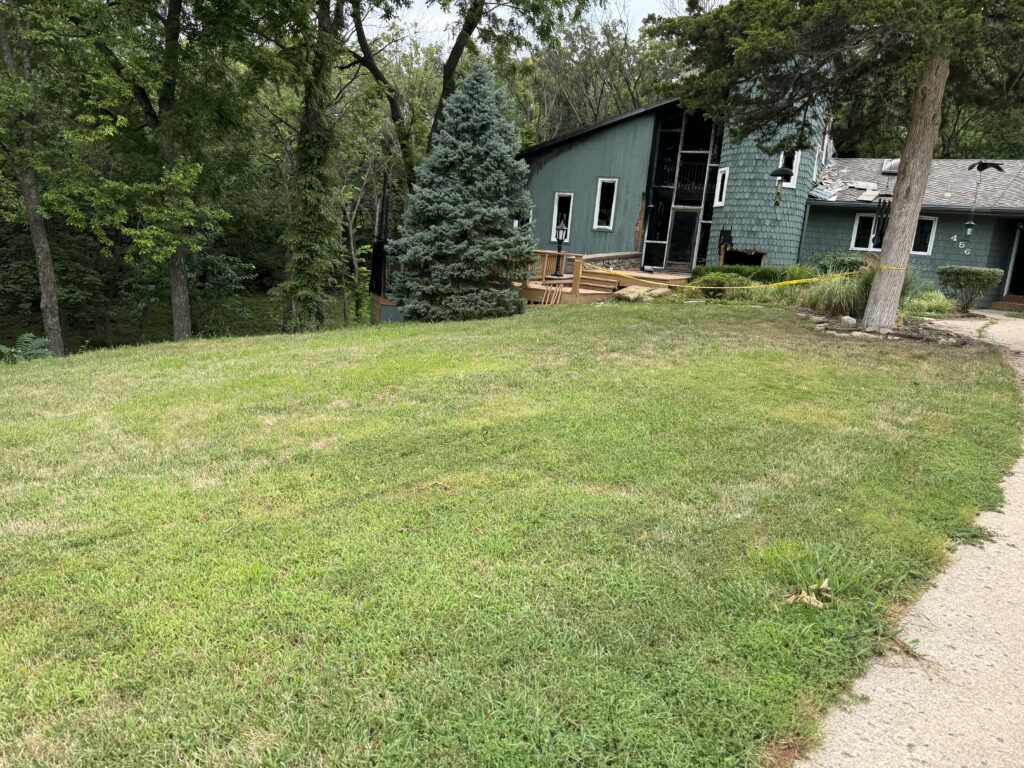 Green yard with manicured lawn, trees, deck, and green house showing visible fire damage.