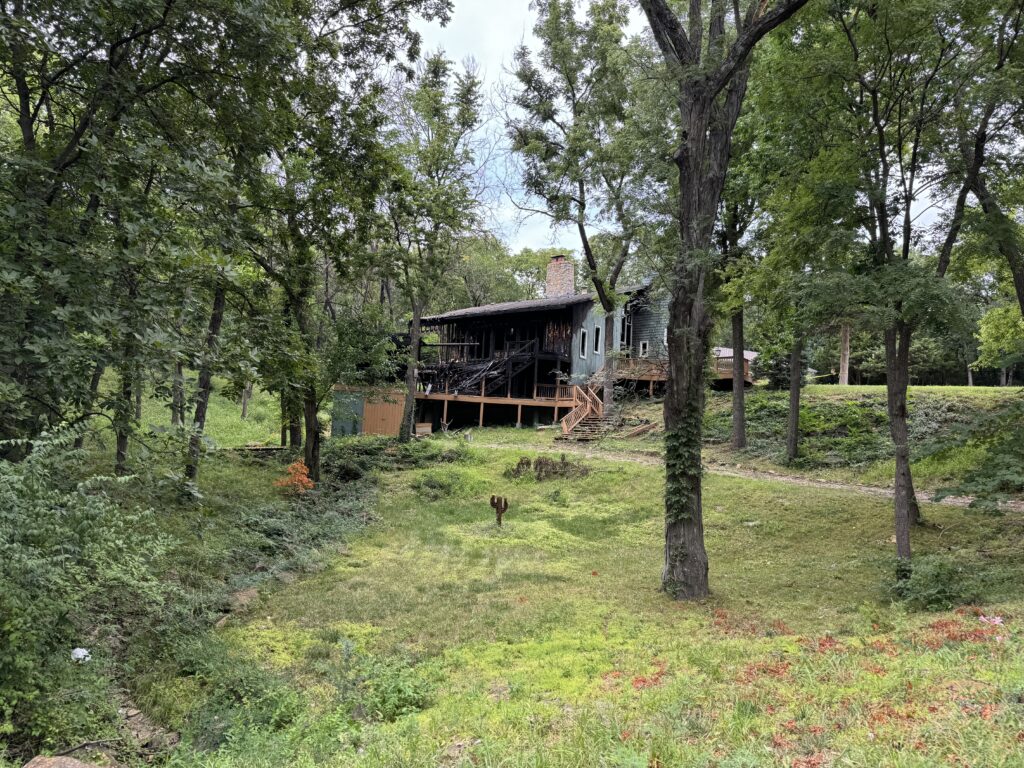 House with fire damage surrounded by trees, grassy lawn, and adjacent small shed with wooden deck.