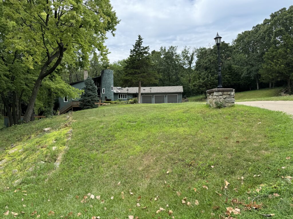 House with trees, grassy lawn, paved driveway, lamp post on stone base, and overcast sky.