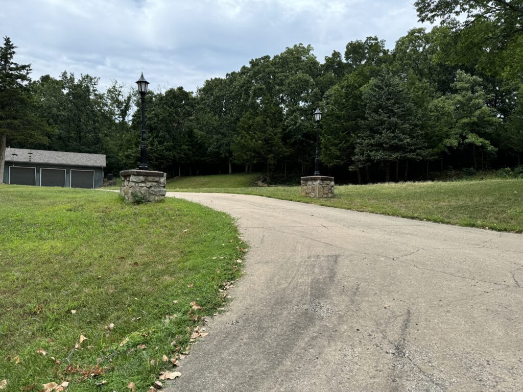 Paved driveway with lampposts leading to a building with garage doors, flanked by dense trees.