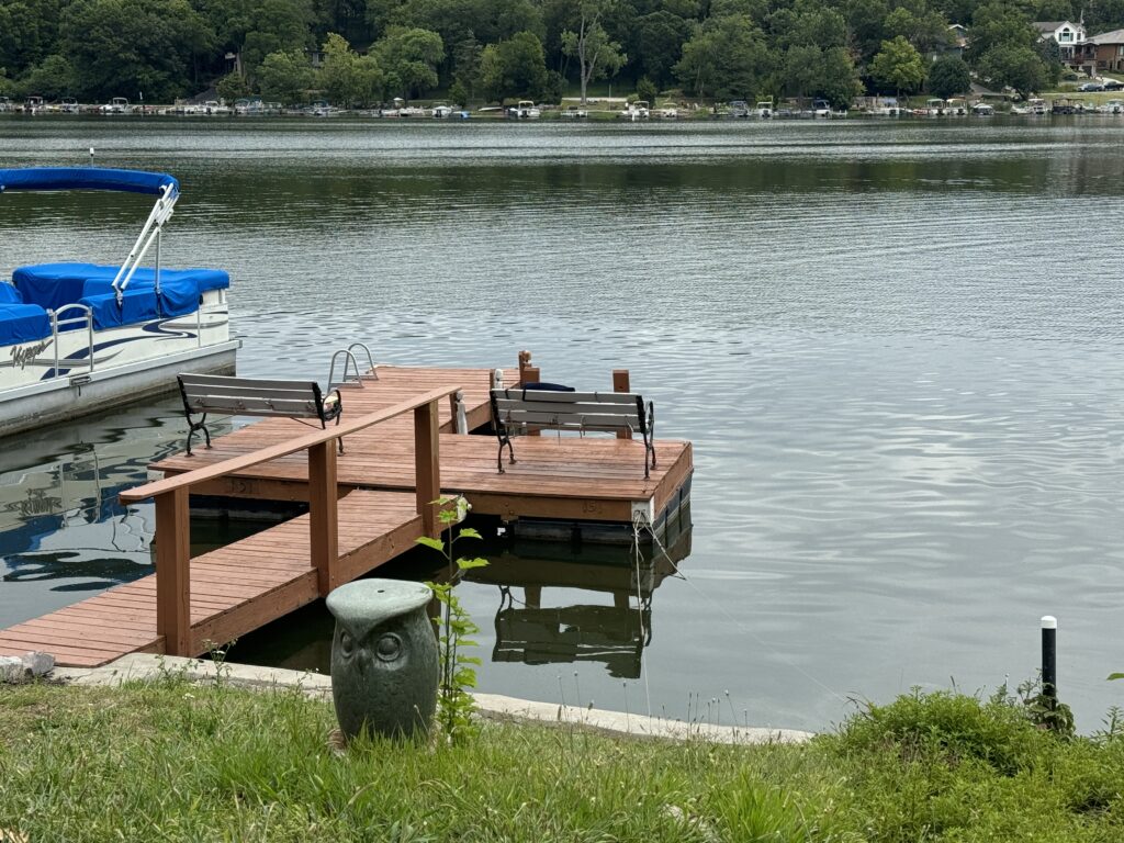 Wooden dock with benches and a pontoon boat on a lake with grassy shore and distant houses.