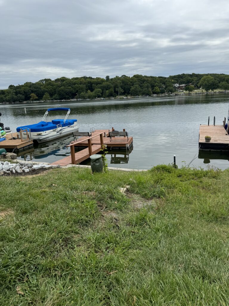 A grassy area leads to docks with boats on a calm lake, surrounded by trees and overcast sky.