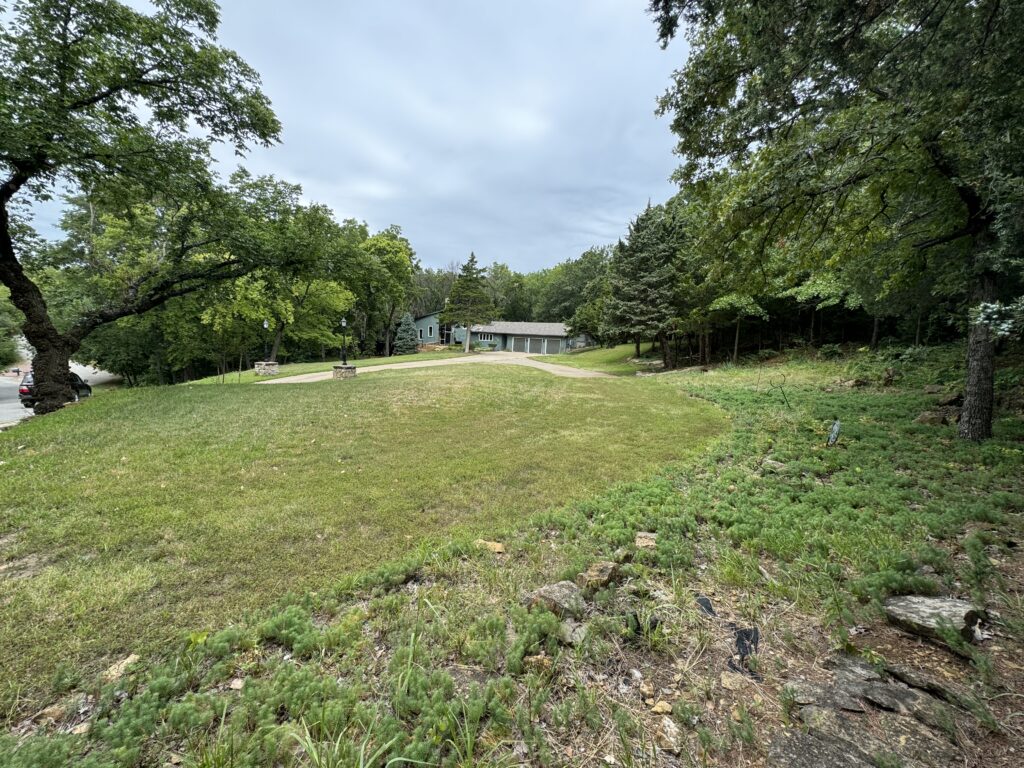 Grassy area with pathway, trees, shrubs, building in background, parked car on left, overcast sky.