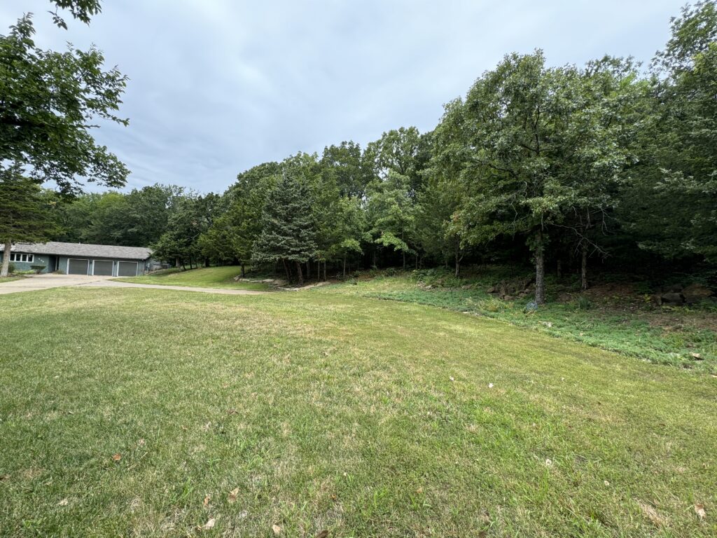 Spacious grassy yard, dense trees to the right, house with three-car garage and driveway to the left.