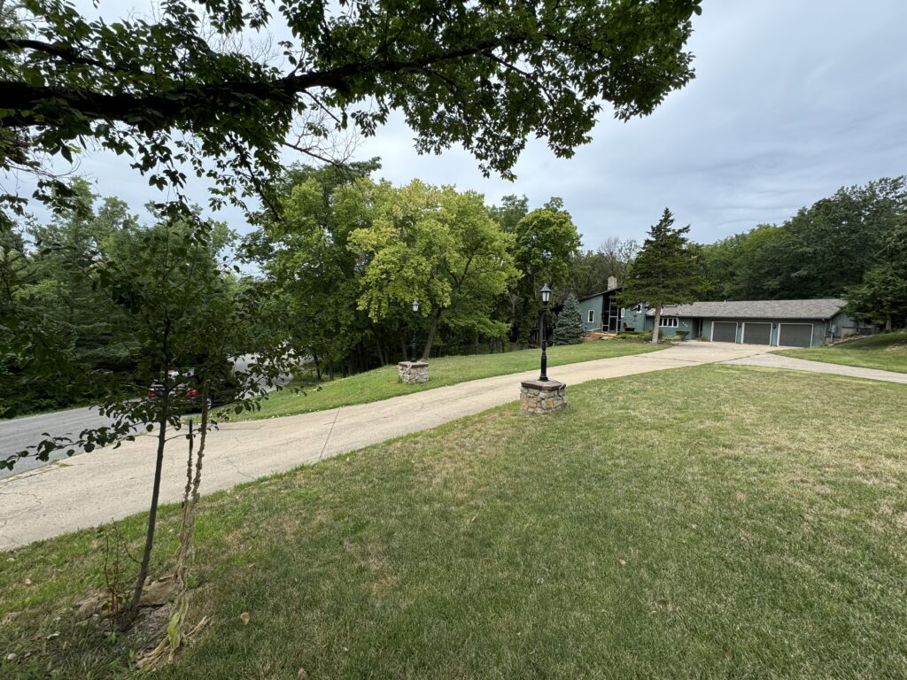 Suburban property with house, multi-car garage, driveway, lamppost, lawn, lush trees, overcast sky.