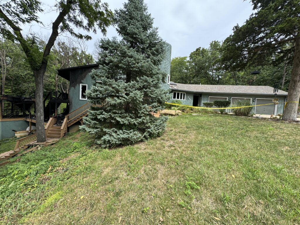 Two-story green house with wraparound deck, wooden stairs, trees, garage, and caution tape on lawn.