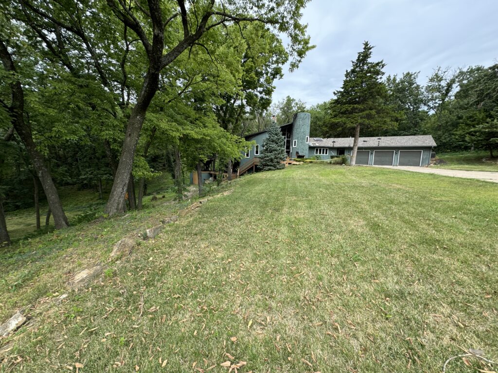 House with multiple levels and cylindrical structure in grassy yard, surrounded by dense trees.