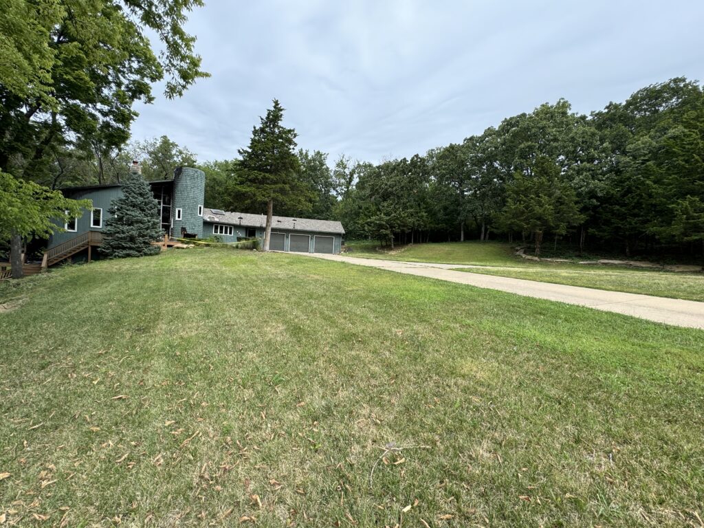 Two-story green house on spacious lawn with paved driveway, grey garage, surrounded by trees, overcast sky.