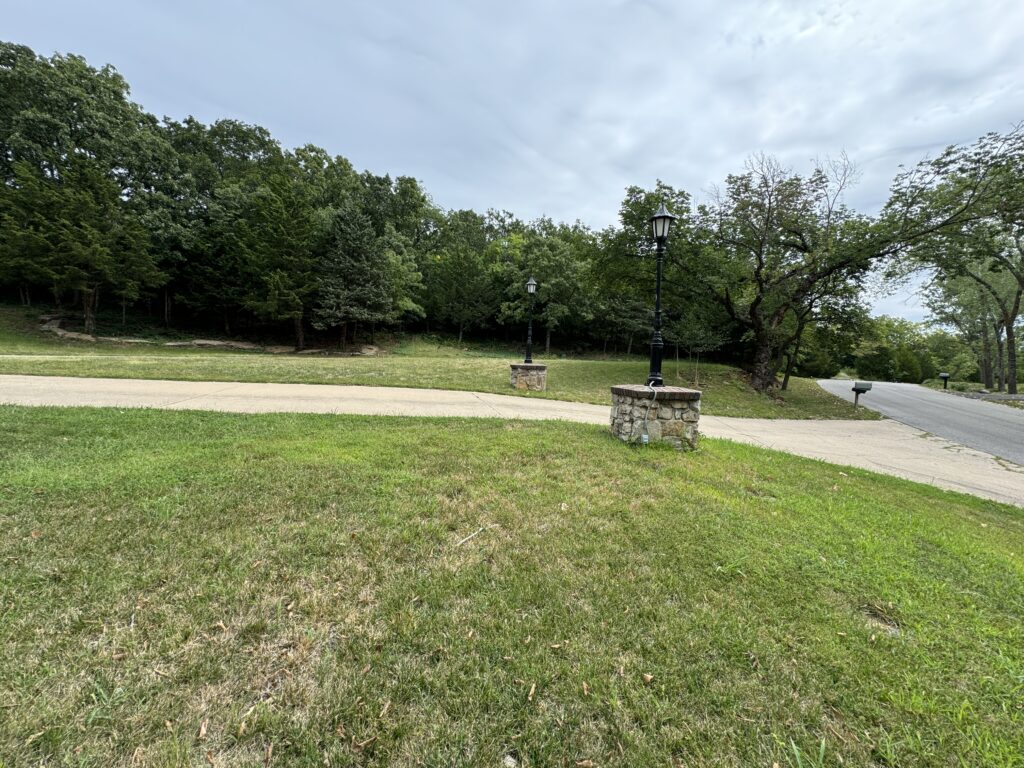 Grassy area with sidewalk, stone lamp posts, paved road, and trees under an overcast sky.