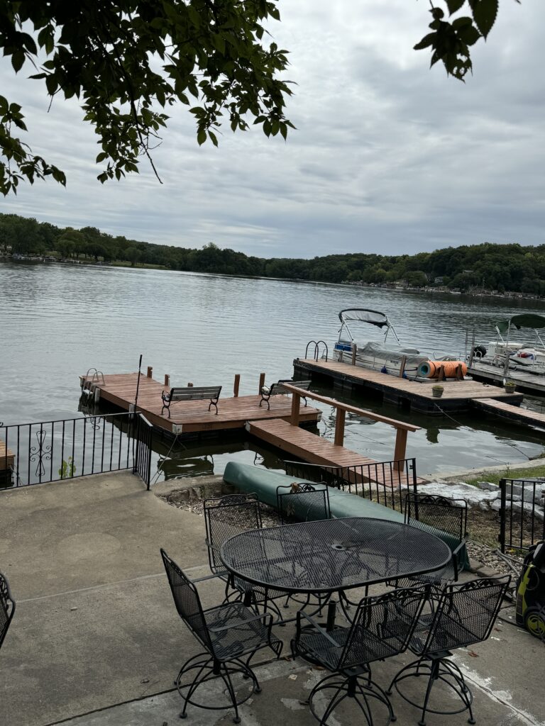 Calm lake view with cloudy sky, surrounded by greenery, featuring an outdoor patio with chairs and docks.