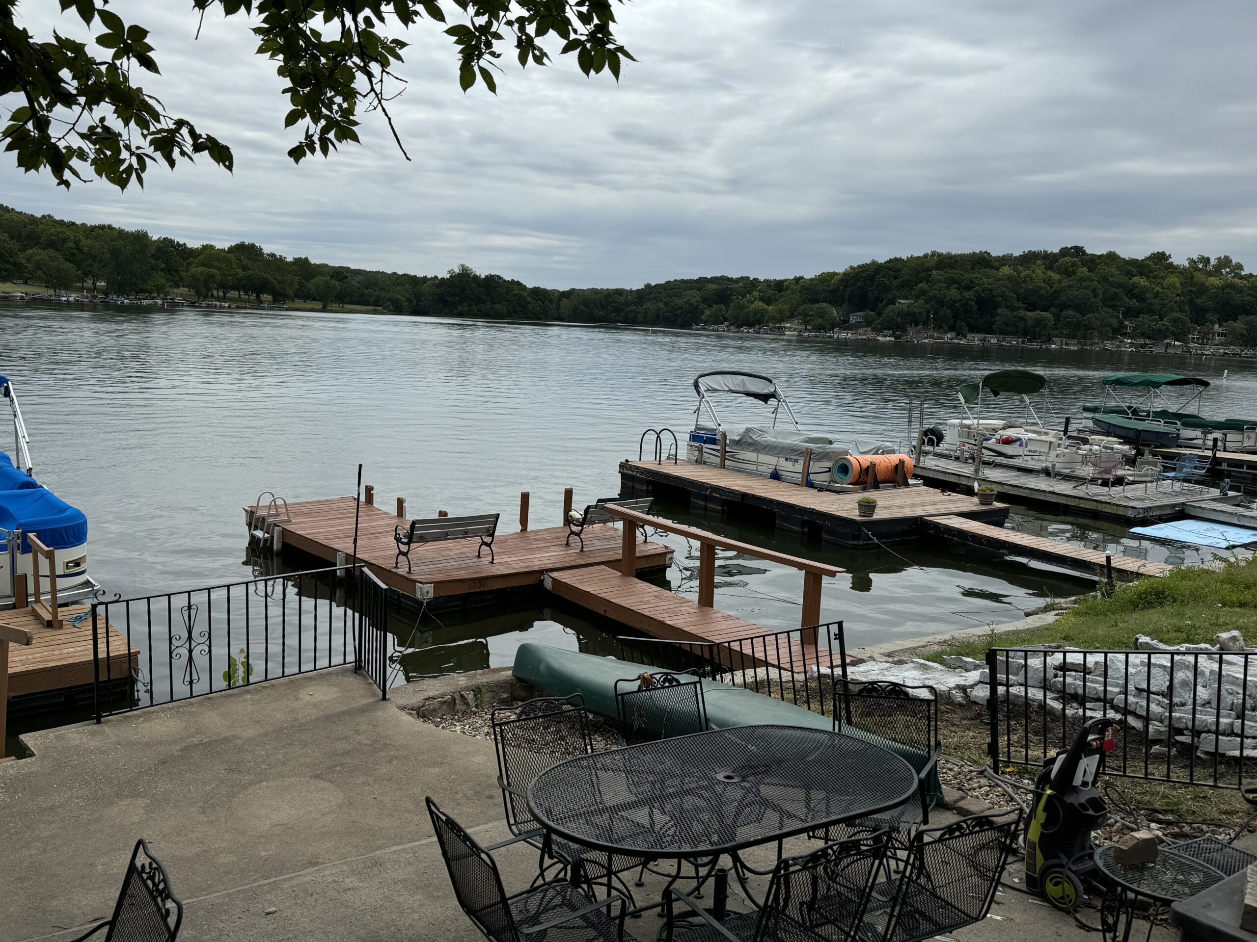 Calm lake with wooden docks, boats tied, patio area with metal furniture, trees, and cloudy sky.