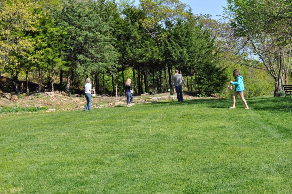 Four people walking in a grassy field near a forest, each carrying a basket. Background features trees.