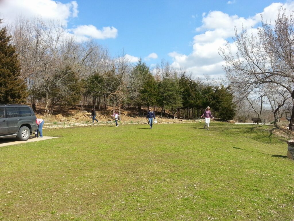 Grassy area with individuals carrying baskets, a parked vehicle, and trees under a partly cloudy sky.